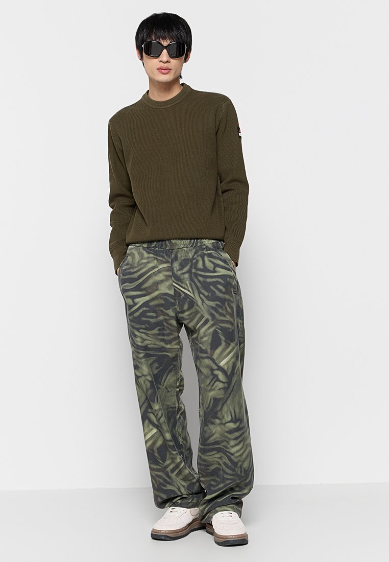 Maglione a coste verde oliva abbinato a pantaloni larghi neri e verdi a fantasia. Sneakers bianche completano l'outfit. Indossati occhiali da sole scuri.