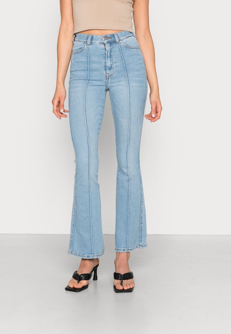Femme portant un jean flare taille haute bleu clair avec coutures frontales, un crop top beige et des sandales à talons noirs avec brides sur les orteils.