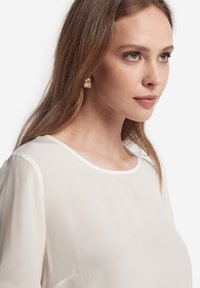 Vit topp med rund halsringning, mjuk textur och korta ärmar. Guldtexturerade örhängen som bärs. Håret flödar löst över axeln.