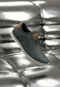 Scarpa in pelle grigia scura con finitura liscia, suola bianca e dettagli color cuoio; presenta una punta arrotondata e un design di allacciatura semplice.
