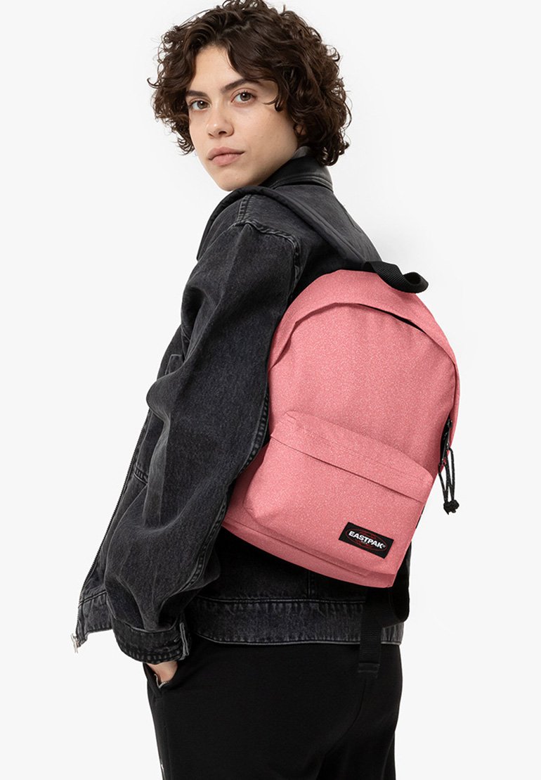 Mochila de tela rosa con superficie texturizada, bolsillo delantero y correas ajustables, llevada sobre un hombro con una chaqueta de mezclilla negra.