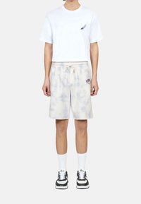 Vit bomulls-T-shirt med en liten fjädergrafik, kombinerad med ljusgrå batikshorts och svarta sneakers med vita accenter och strumpor.