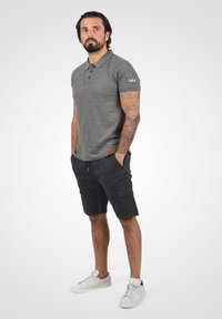 Tricou polo gri cu trei nasturi maronii, asociat cu pantaloni scurți cargo bleumarin. Material texturat și detaliu cu logo pe mânecă. Adidași albi.