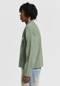 Groene gebreide trui met lange mouwen en een textuurstof. Heeft een ontspannen pasvorm en geribbelde manchetten. Gecombineerd met lichtblauwe denim.