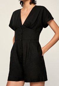 Czarny krótki kombinezon z dekoltem w kształcie litery V, krótkimi rękawami, pięcioma guzikami z przodu oraz bocznymi kieszeniami, charakteryzujący się luźnym krojem i miękką teksturą.