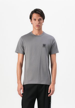 Young man standing, wearing a plain grey t-shirt with a small logo on the chest and black pants against a white background.