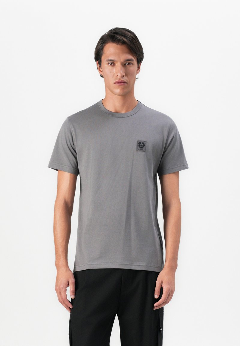 Young man standing, wearing a plain grey t-shirt with a small logo on the chest and black pants against a white background.
