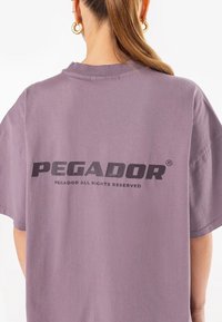 Ljubičasta oversized majica, pamučni materijal, okrugli izrez, kratki rukavi, s bold crnim natpisom "PEGADOR" na leđima, zajedno s logotipom i detaljima o autorskim pravima.