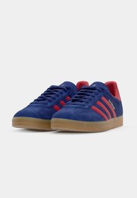 Blå ruskind sneakers med røde striber, gummirubber såler og "GAZELLE" branding på siden.