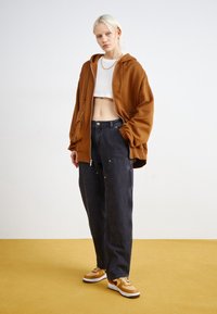 Överdimensionerad brun huvtröja, vit kort topp, mörka högmidjade jeans med fickor, och orange sneakers på ett gult golv. Enkel bakgrund.