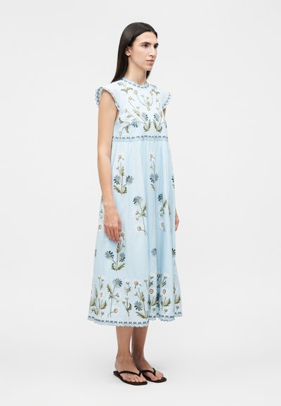 Γυναίκα που στέκεται, φοράει ένα γαλάζιο μίντι φόρεμα με κεντήματα λουλουδιών και κυματοειδείς μανσέτες, συνδυασμένο με μαύρα σαγιονάρες.