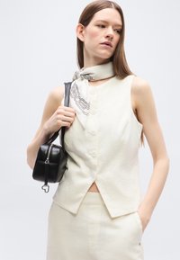 Γυναίκα που φοράει μπεζ τοπ χωρίς μανίκια με κουμπιά και παντελόνι, λευκό μαντήλι με σχέδια, κρατώντας μαύρη ορθογώνια τσάντα στον ώμο.