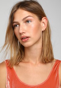 Modella con capelli castani dritti alla spalla, che indossa una canotta di velluto corallo. Orecchini minimalisti a cerchio d'oro adornano le orecchie. Illuminazione soffusa.
