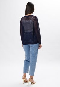 Dunkelblaues Stricktop mit einem Netzmuster, ausgestattet mit langen Ärmeln und einem rundem Ausschnitt; kombiniert mit hellblauen Jeans und goldenen Absätzen.