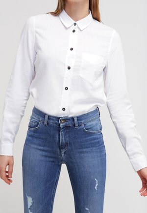 Persoon die een wit overhemd met zwarte knopen draagt, ingestopt in een blauwe versleten spijkerbroek, tegen een effen achtergrond.