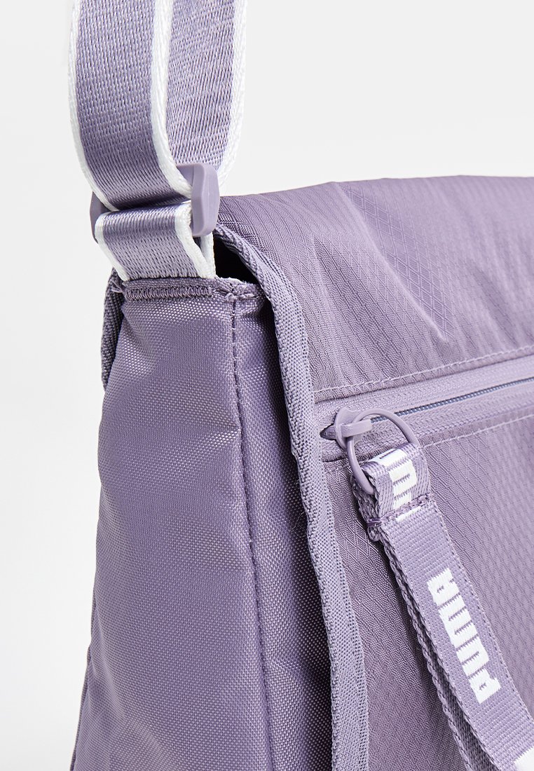 Bolsa textil púrpura con superficie texturizada, correa ajustable blanca y marca PUMA. Incluye un bolsillo lateral con cremallera para almacenamiento.