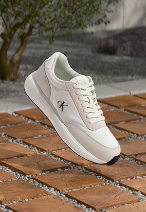 Bílá teniska Calvin Klein s logem na boku, bílými tkaničkami a robustní podrážkou položená na dřevěných čtvercových dlaždicích s kamennými mezerami venku.