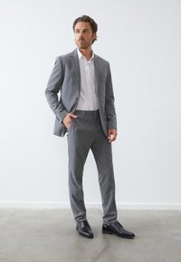 Grau maßgeschneiderter Anzug mit schmalem Schnitt, strukturiertem Stoff und Zwei-Knopf-Verschluss. Ein weißes Dress-Shirt und schwarze Lederschuhe runden den Look ab.