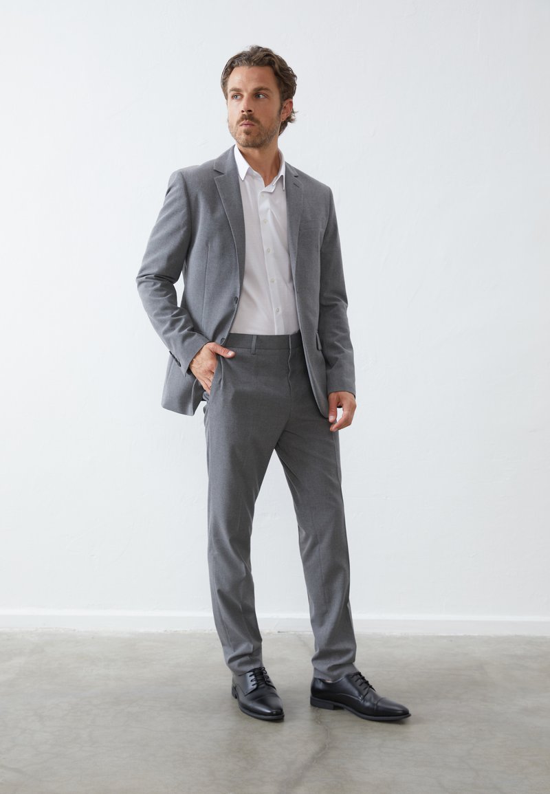 Grau maßgeschneiderter Anzug mit schmalem Schnitt, strukturiertem Stoff und Zwei-Knopf-Verschluss. Ein weißes Dress-Shirt und schwarze Lederschuhe runden den Look ab.