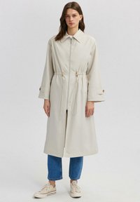 Trench coat bej deschis cu fermoar în față, talie cu șnur și mâneci late. Asortat cu blugi albaștri cu manșetă și adidași albi.
