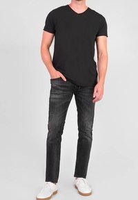 Hombre vestido con camiseta negra de cuello en V, jeans negros desgastados y zapatillas blancas, de pie con una mano en el bolsillo frente a un fondo sencillo.