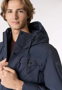 Giacca imbottita blu navy con cappuccio, bottoni a scatto e taschino sul petto. Tessuto testurizzato con lacci regolabili per una vestibilità ottimale.