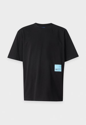 Jeune homme debout, une main dans la poche, portant un t-shirt noir oversize avec un petit patch bleu et un pantalon noir ample avec des baskets noires.