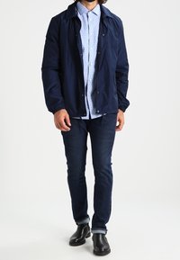Chaqueta de nailon navy sobre una camisa de lunares de manga corta en azul claro, combinada con jeans oscuros y botas negras de tobillo.