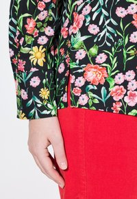 Blusa negra con estampado floral en tonos vibrantes de rosa, amarillo y verde; combinada con pantalones de pana rojos. Mano visible descansando sobre la costura.