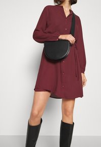 Bordeaux, langærmet skjorte-kjole med en struktureret mønster og et bånddetalje i taljen, accessoriseret med en sort crossbody-taske og støvler til knæene.