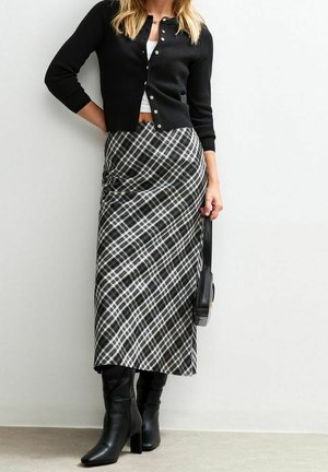 Kvinde iført sort cardigan, hvid crop top, sort-hvid ternet nederdel, sorte støvler med hæl og holder en sort håndtaske.