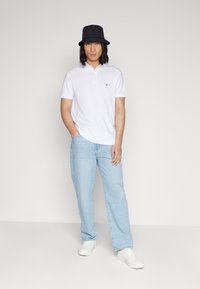 Polo bianco, jeans azzurri chiari e sneakers bianche. Cappello a secchiello nero. Modello in piedi contro uno sfondo neutro con le mani in tasca.