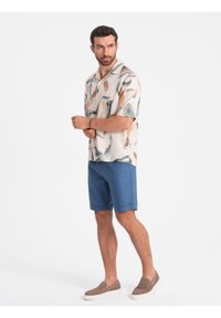 Chemise en lin à manches courtes avec un fond beige et des motifs abstraits gris, marron et orange, associée à un short bleu et des chaussures à enfiler.