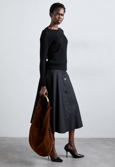 Pull noir à côtes, jupe noire taille haute avec des boutons dorés, sac en daim brun et escarpins noirs sur un sol gris.