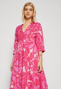 Lång, rosa klänning med blommönster, med V-ringning, trekvartslånga ärmar, knappproppar och plisserad kjol. Slät tygtextur.