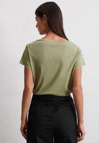 Olivgrünes T-Shirt mit kurzen Ärmeln und weitem Halsausschnitt, kombiniert mit schwarzen hochtaillierten Hosen mit Taschen auf der Rückseite und Knopf-Akzenten.