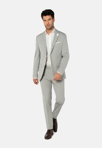 Costume gris clair avec une coupe ajustée, veste à un seul bouton, revers en notch, et poche carré blanche. Des chaussures habillées marron complètent le look.