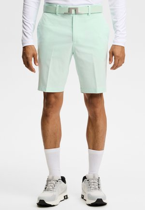 Mand iført lyse mintgrønne skræddersyede shorts med matchende bælte, hvid langærmet top, hvide sokker og hvide sneakers.