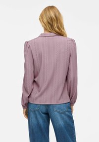 Lilla strukturert bluse med krage, puffete ermer og knappelukking, med dekorative hullmønstre nedover stoffet.