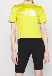 Żółta koszulka sportowa z krótkim rękawem i białym logo, zestawiona z czarnymi dopasowanymi szortami rowerowymi. Gładka tekstura i luźny krój.