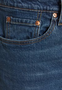 Tissu en denim bleu foncé avec une texture lisse. Comprend une petite poche et des rivets en cuivre. Des coutures jaunes soulignent les coutures.