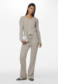 Striped loungewear-set i beige og svart, med en knapped V-hals topp og bukser med snor. Modell holder en sølvveske.