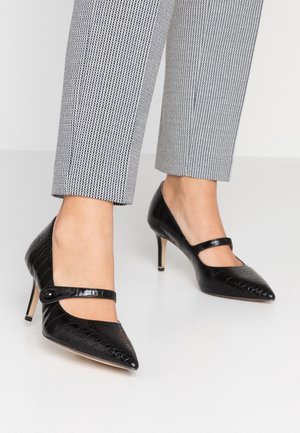 Fødder iført sorte laklæder Mary Jane-pumps med spidse tå, kombineret med slanke, grå ternede ankelkorte bukser på en hvid gulv.