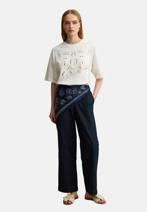 Mujer que lleva un top blanco de manga corta con aberturas, cinturón de pañuelo azul marino con estampado, pantalones anchos oscuros y sandalias beige tejidos sin cordones.