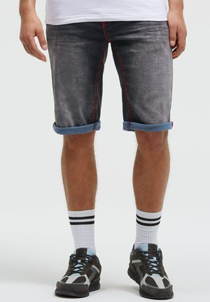 Mann trägt hochgekrempelte dunkelgraue Jeansshorts mit roter Naht, weiße Socken mit schwarzen Streifen und schwarze und graue Sneakers.