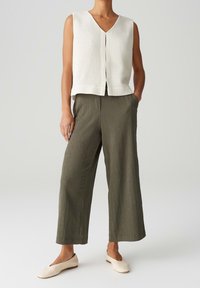 Bílý texturovaný bezramínkový top s výstřihem do V, doplněný olivově zelenými širokými kalhotami a béžovými plochými botami, na světlém pozadí.