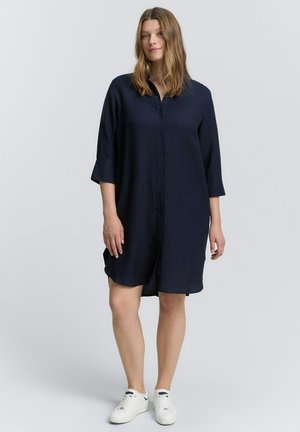 Marineblaue Knopfhemdkleid aus leichtem Stoff, mit Dreiviertelärmeln, seitlichen Schlitzen und einem lockeren Schnitt. Getragen mit weißen Sneakers.