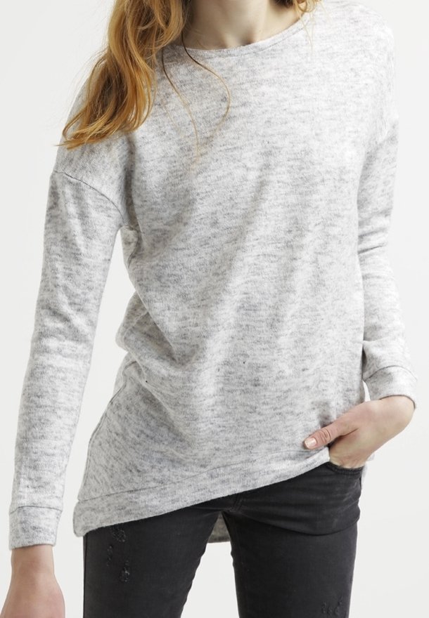 Femme portant un pull à manches longues gris clair et un pantalon noir, une main dans la poche, debout devant un fond uni.