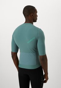 Koszulka sportowa z krótkim rękawem w kolorze teal z heksagonalną teksturą, z zielonym logo i dopasowanym krojem.