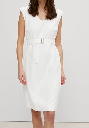 Frau trägt ein ärmelloses, weißes, knielanges Kleid mit V-Ausschnitt und passendem Gürtel mit rechteckiger Schnalle.